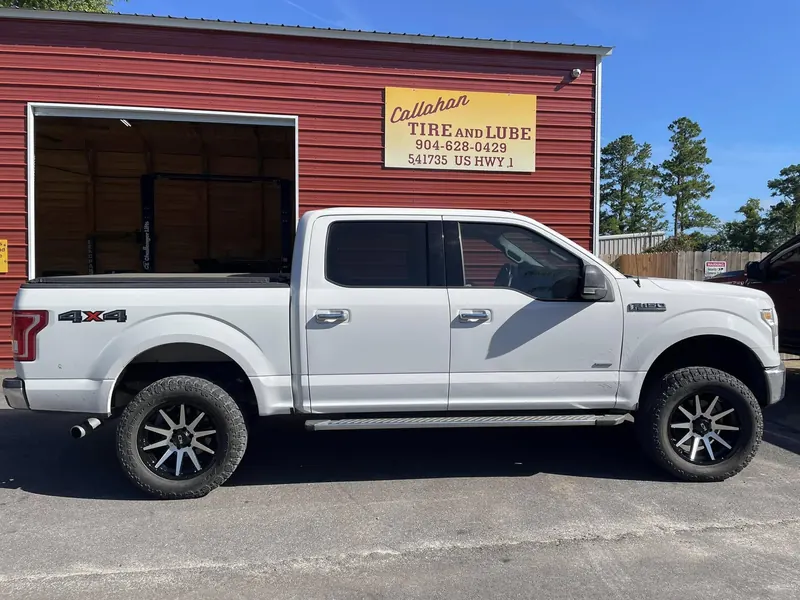White Ford F-150 4x4 on black wheels under the Callahan Tire and Lube sign, an open bay with a lift behind it